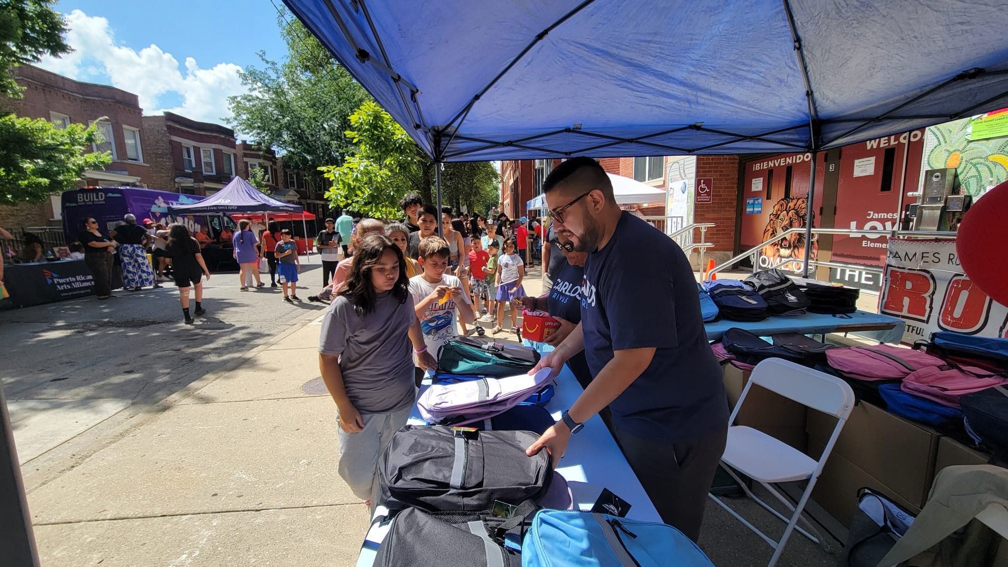 Tents line a street. A man organizes backpacks while students line up to receive them.