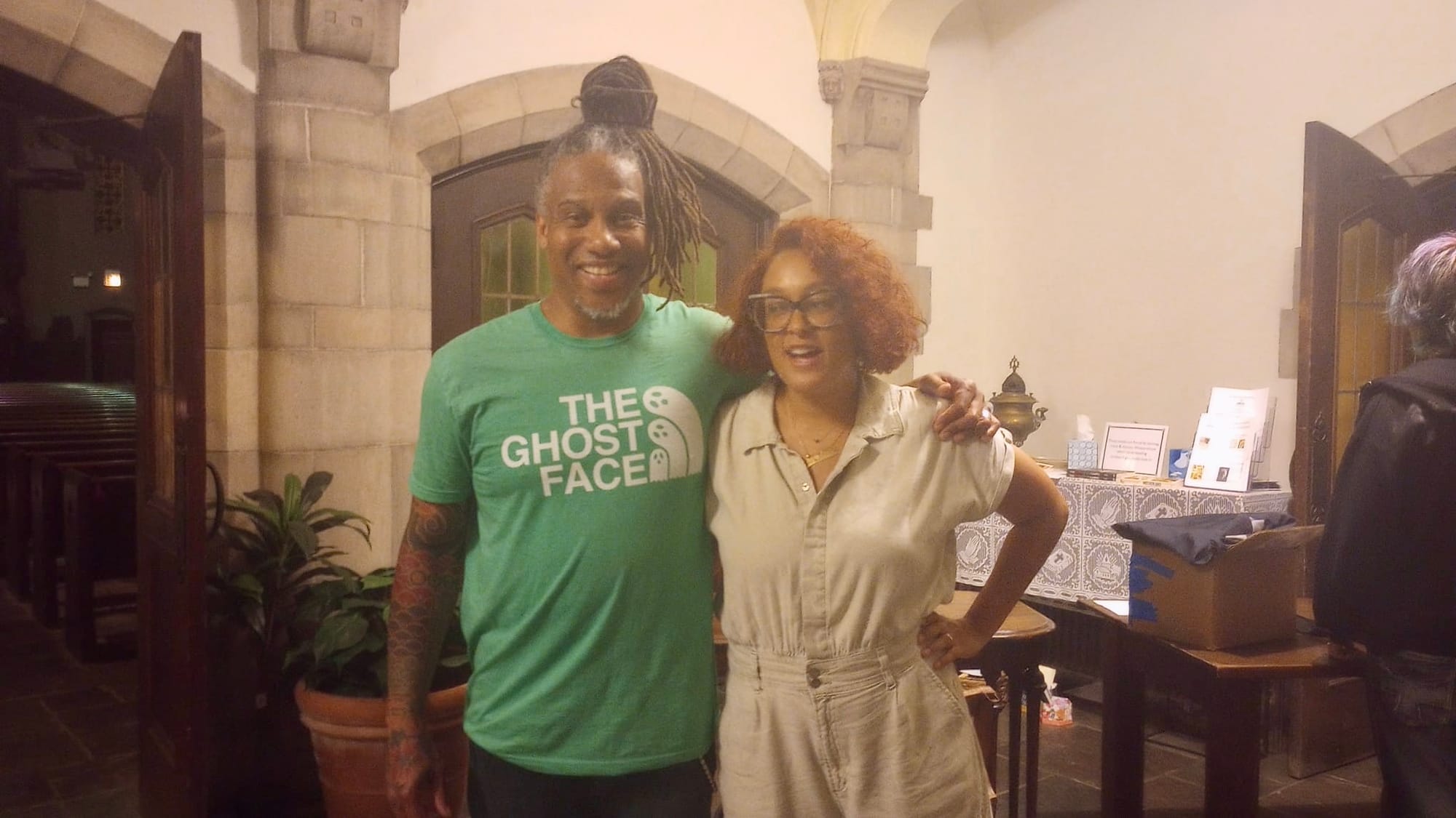 David Stovall, a tall man with dreadlocked hair in a green t-shirt stands next to Eve Ewing, a curly-haired woman in a khaki dress.