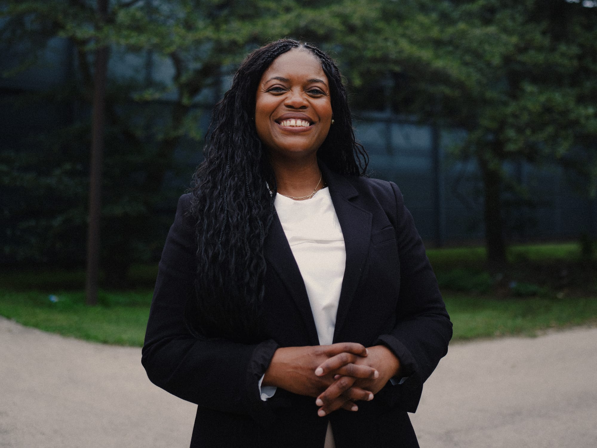 A long-haired Black woman smiles broadly while standing, hands folded, in front of a school building.
