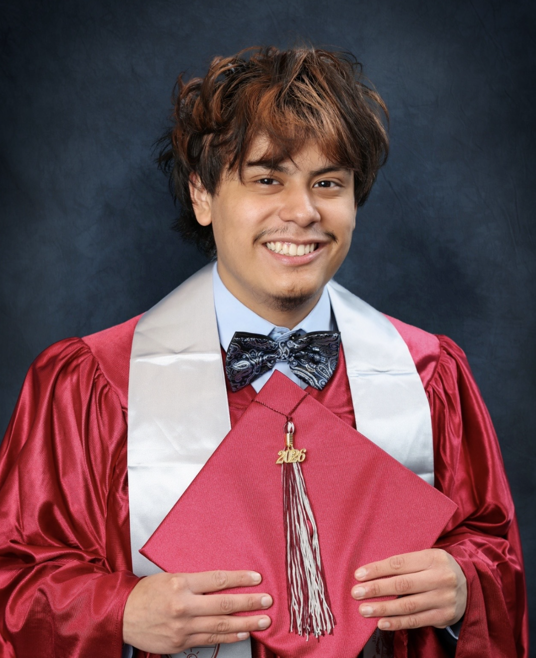 A young man in a red graduation gown with a white stole holds a red mortarboard with a red and sliver 2026 tassel.