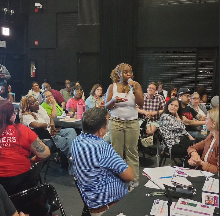 A young woman holds a cordless mic while speaking to a crowd of people in a black-box theater.