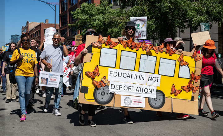 Six people hold up a large piece of cardboard painted with a yellow school bus and a sign saying "Education Not Deportation."