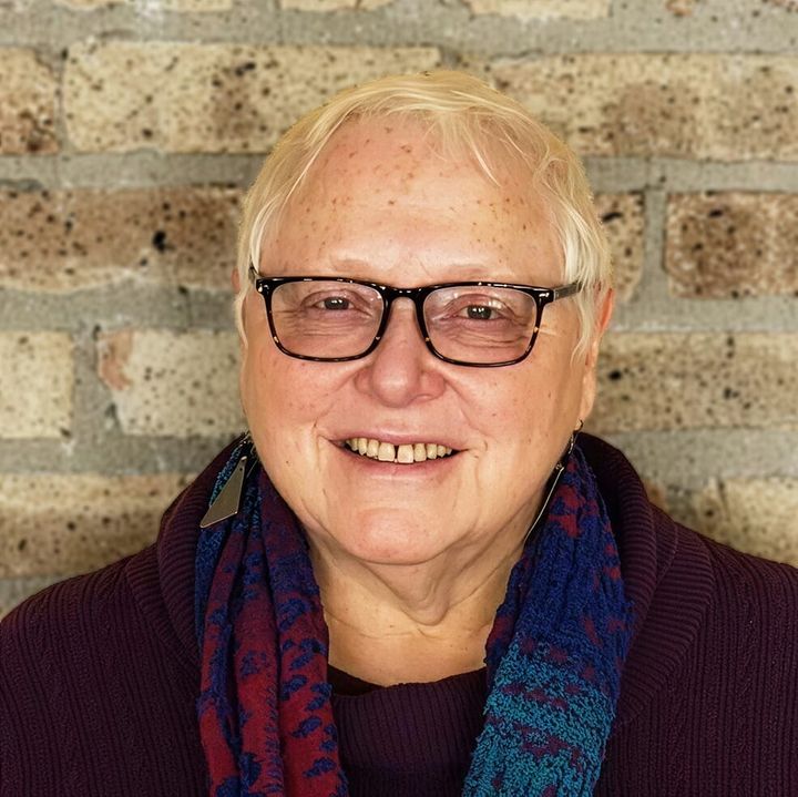 The photo shows a woman with short blond hair and glasses, wearing a maroon and blue scarf and a maroon sweater.