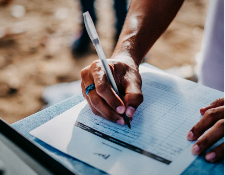 The photo centers on a right hand holding a pen and signing a sheet of paper with columns.