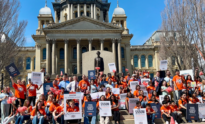 People wearing orange shirts and holding signs asking for $550M for schools stand on the capitol steps in Springfield.