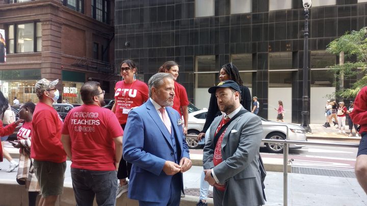 A small group of people in red t-shirts stand in the background. Foreground, a man in a light blue suit and pink tie speaks with a man in a baseball hat.