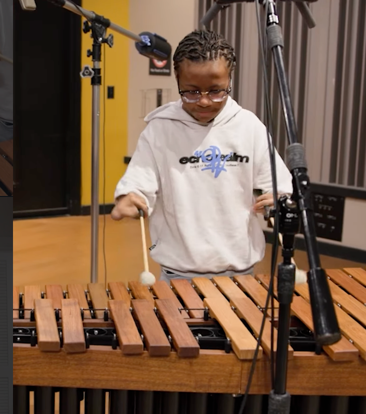 A young man with braids and glasses plays the marimba. Microphones stand in front of him and to his right.