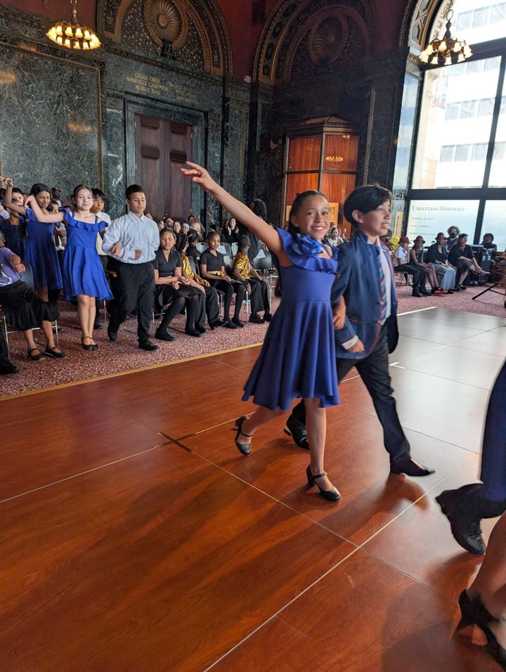 A young girl in a blue, knee-length dress strides a cross a dance floor with her right arm outstretched, accompanied by her dance partner, a young boy.