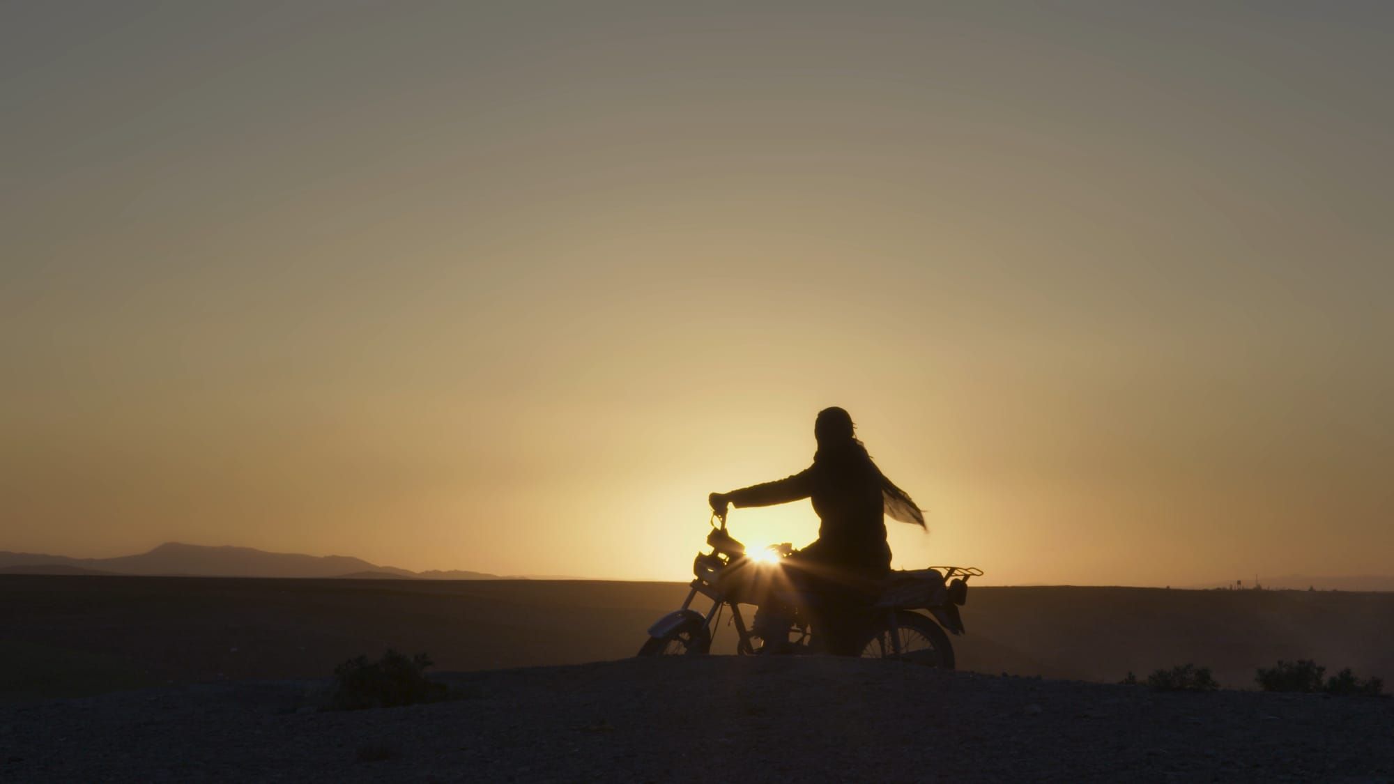 A person riding a motorbike silhouette with a flat horizon and clear sky in the background. Still from the movie CUTTING THROUGH ROCKS.