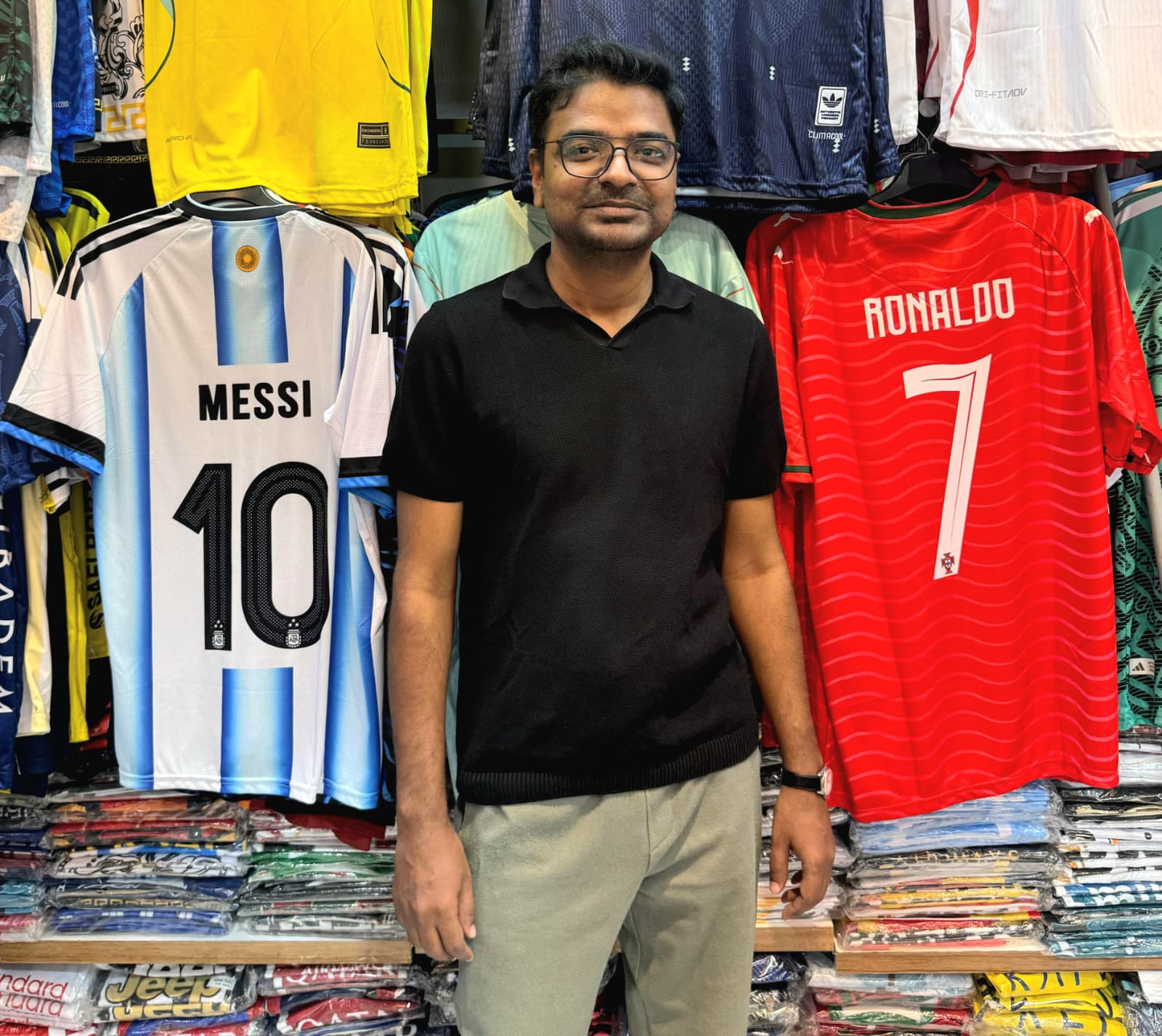 Prof Saifuddin, a man wearing a black shirt and khaki pants, in between Messi and Ronaldo football jerseys in a clothing store.