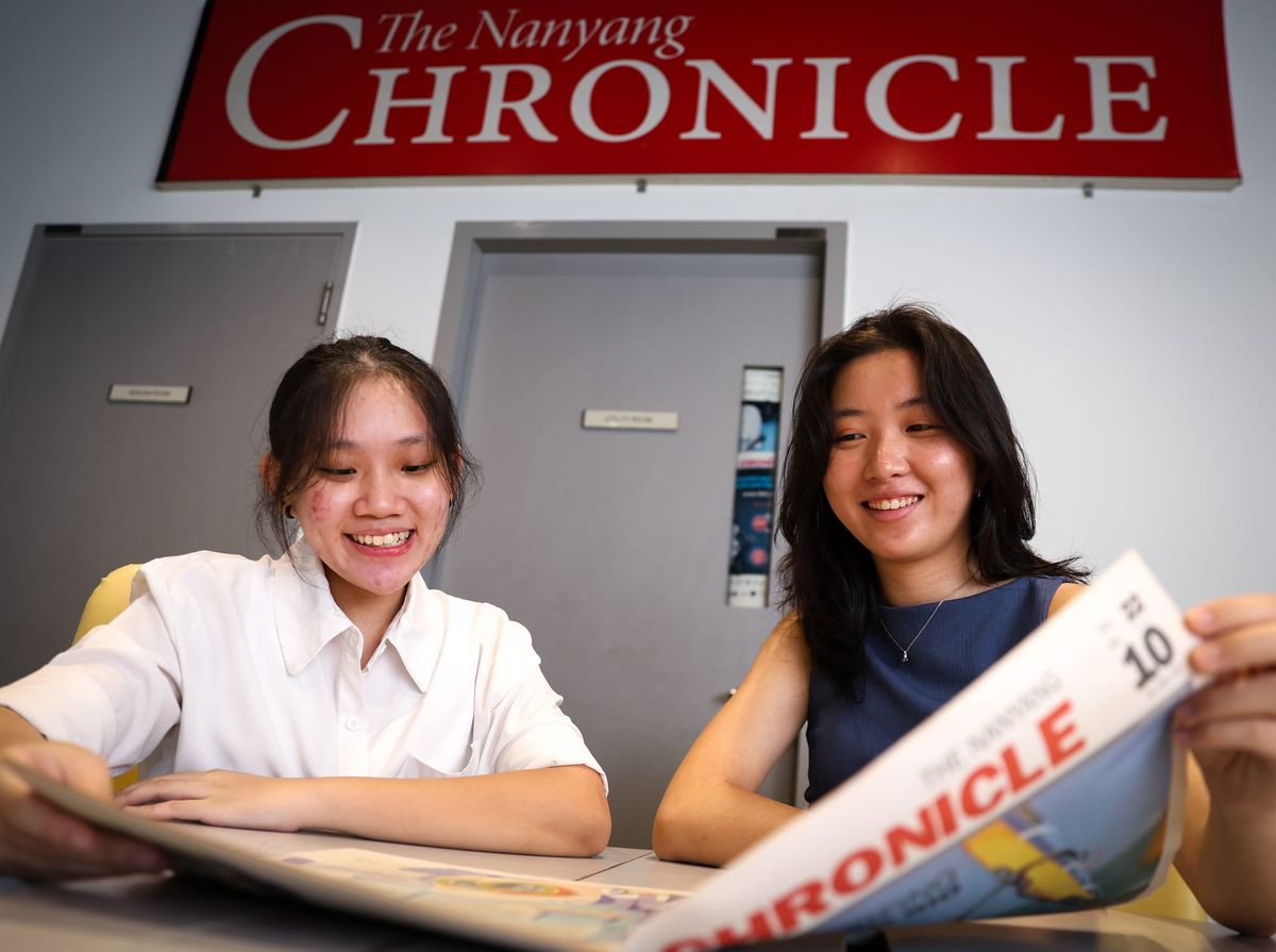 Two girls sitting, smiling and looking at an old print copy of the Nanyang Chronicle. A sign with the words "The Nanyang Chronicle" is displayed on the wall behind them.