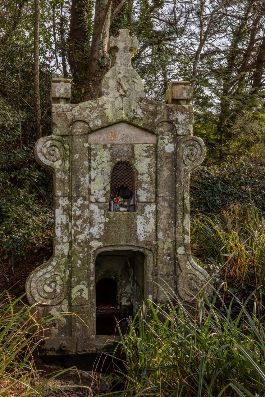 Fontaine gothique en pierre avec croix, nichée dans la végétation au bord de l'eau