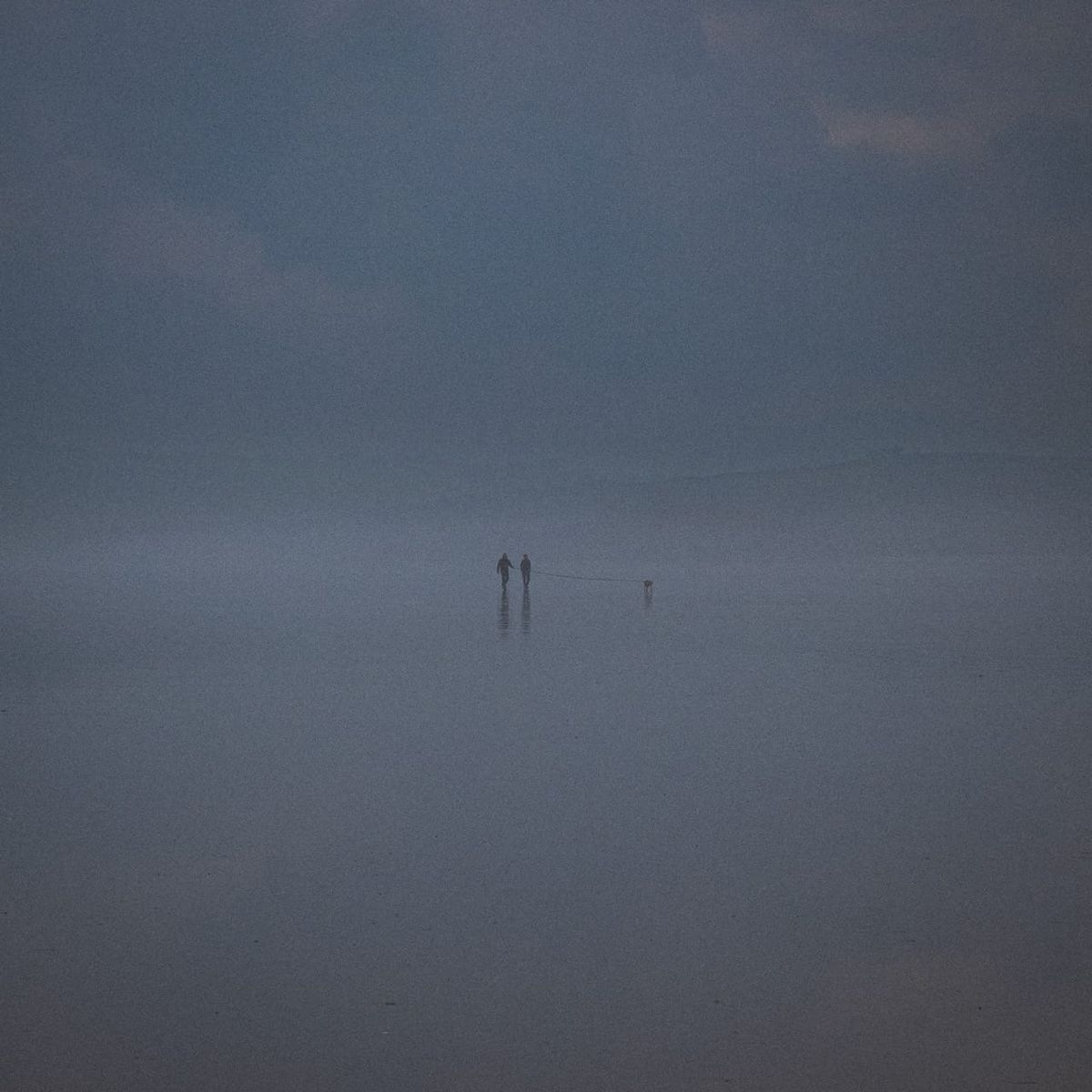 Deux silhouettes marchent avec un chien sur le sable miroir à marée basse dans la brume à Sainte-Anne-la-Palud en Finistère.
