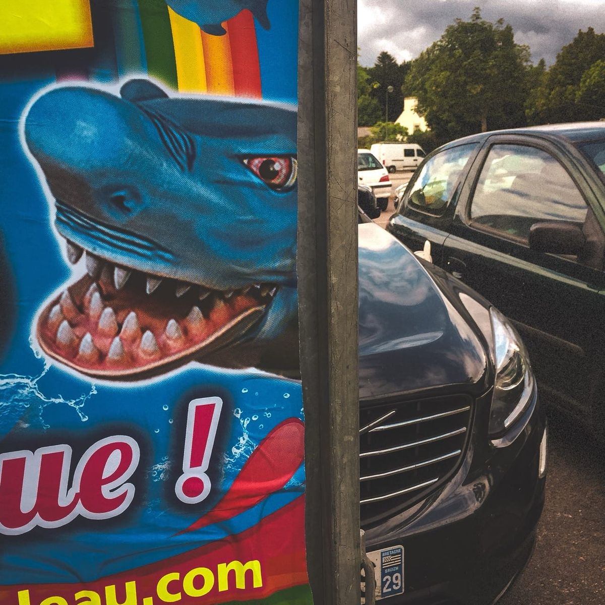 Affiche de requin souriant face à une voiture noire sur un parking à Fouesnant, formes visuelles similaires