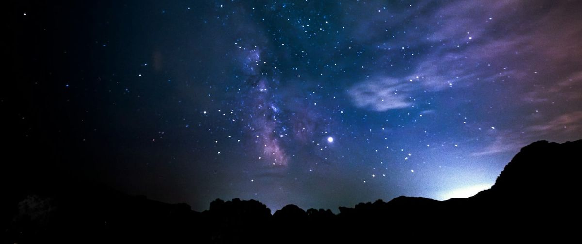 A starry night sky with whisps of blue and purple. The sun peaks out from behind a mountain range.