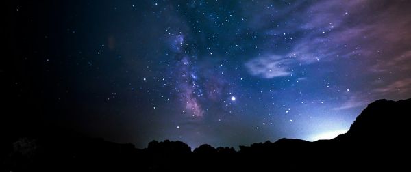 A starry night sky with whisps of blue and purple. The sun peaks out from behind a mountain range.