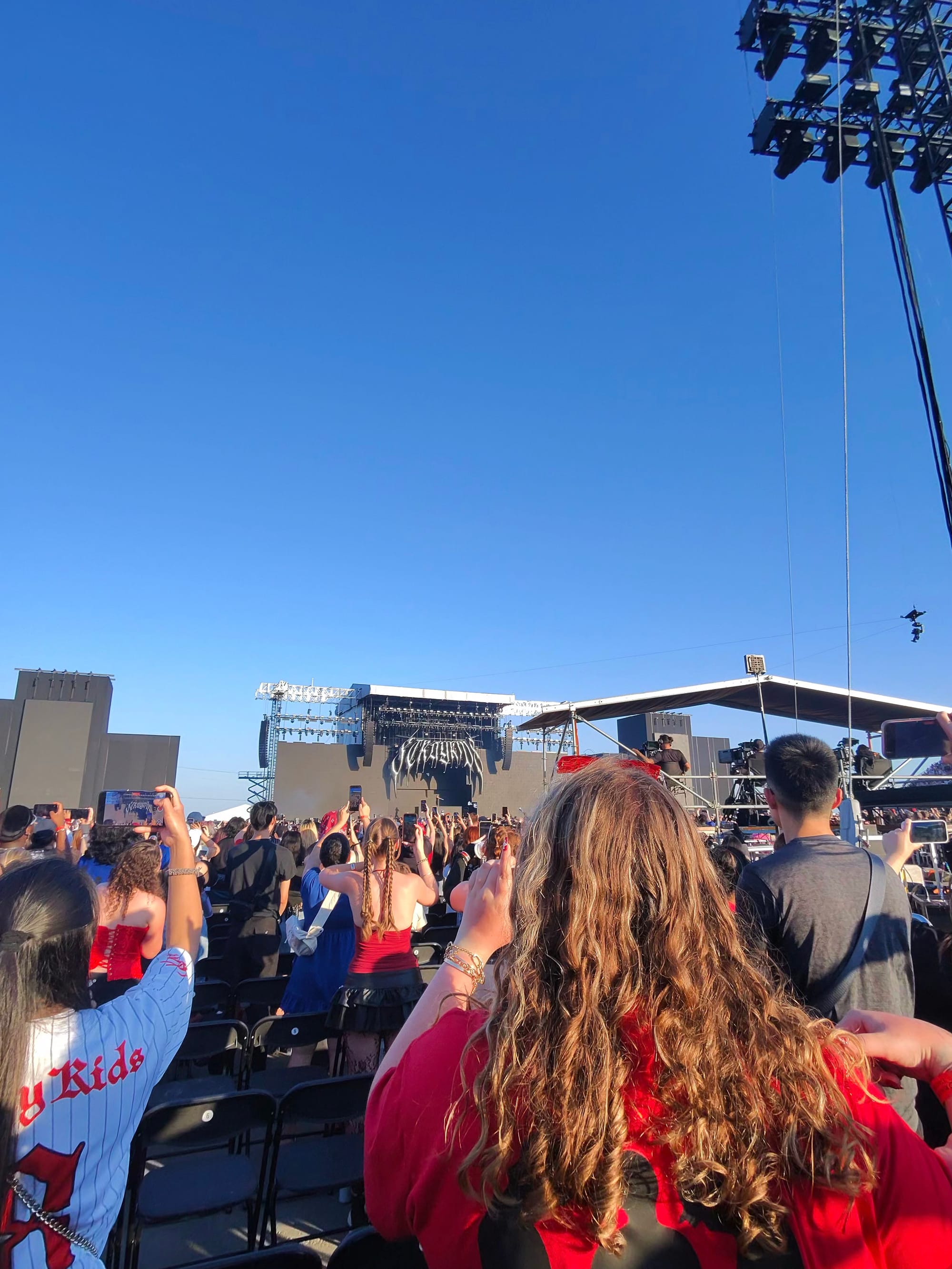  Fans in a crowd in front of a stage; a tent covered podium features videographers to the left of the screen