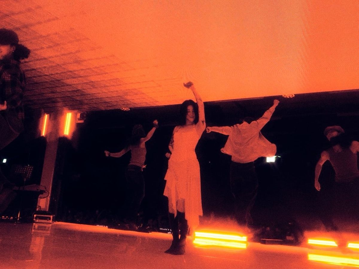 Yerin Baek on stage, revelling in the performance with her dancers. She's in a white dress, awash in orange light.