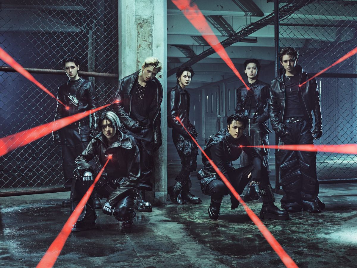 Enhypen members in black backed by chained fences in a concrete parking complex, shining red laser pointers towards the camera 