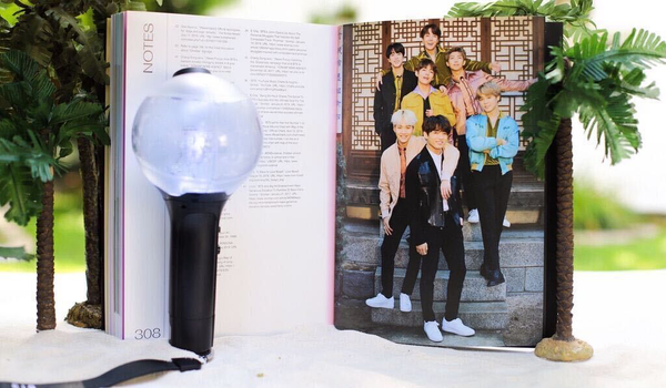 A book open in front of small palm trees, with an Army bomb in front of it. The pages opened are "Notes" bibliography on the left side, and a picture of BTS on the right. 