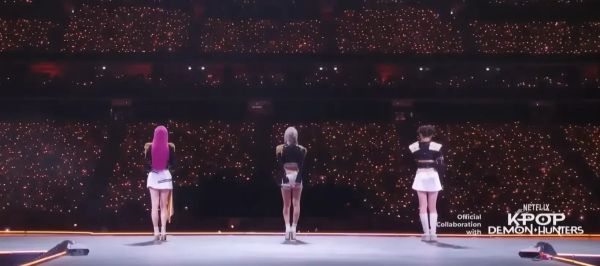 BabyMonster facing away from the camera on-stage, performing K-Pop Demon Hunters at MAMA with the words "Official Collaboration with NETFLIX K-POP DEMON HUNTERS"