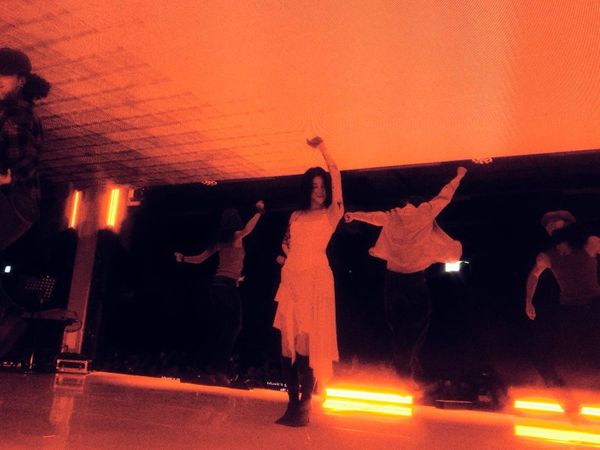 Yerin Baek on stage, revelling in the performance with her dancers. She's in a white dress, awash in orange light.