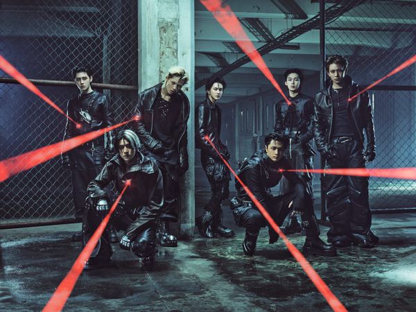 Enhypen members in black backed by chained fences in a concrete parking complex, shining red laser pointers towards the camera 