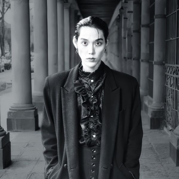 Black-and-white photo of El Capitxn staring down the camera, surrounded by columns in a jacket and ruffled shirt