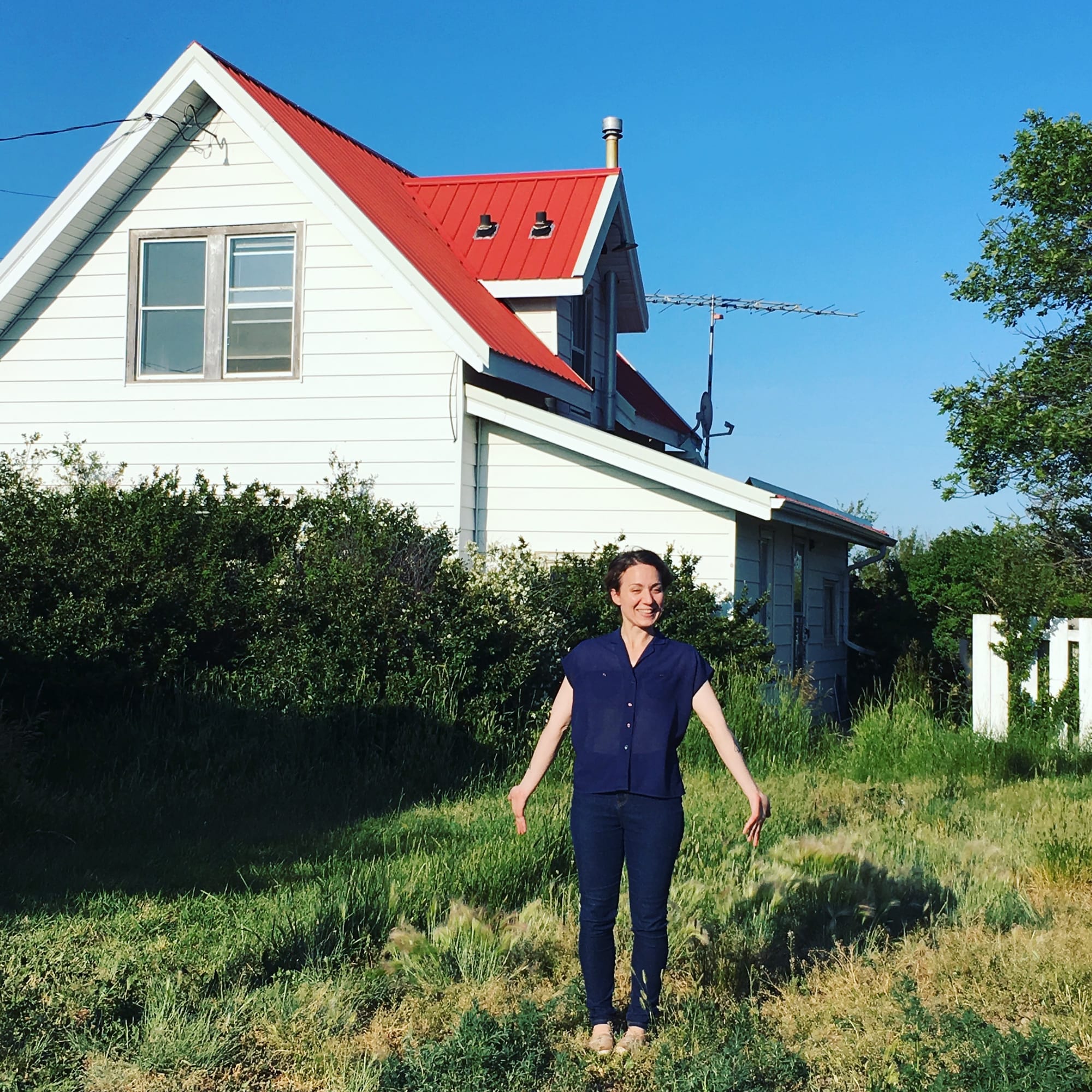 A woman standing in front of an old white farmhouse with a red metal roof. The grass is overgrown.