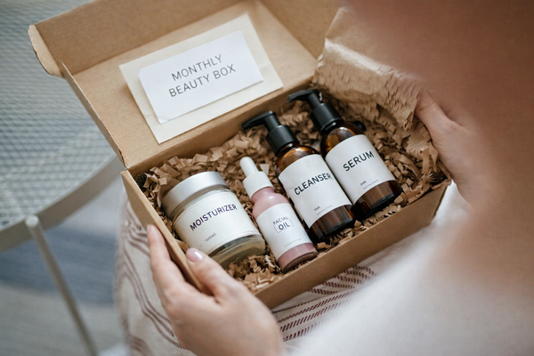 A person holding an open cardboard subscription box filled with neatly arranged skincare products, including bottles and jars, cushioned with packing paper.