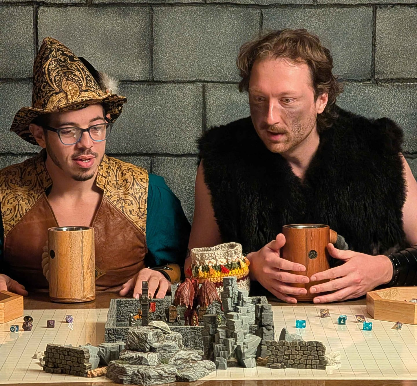 Colton and Wyatt sitting at a special game table, dressing in fantasy outfits holding wooden mugs and staring at 3D models and terrain