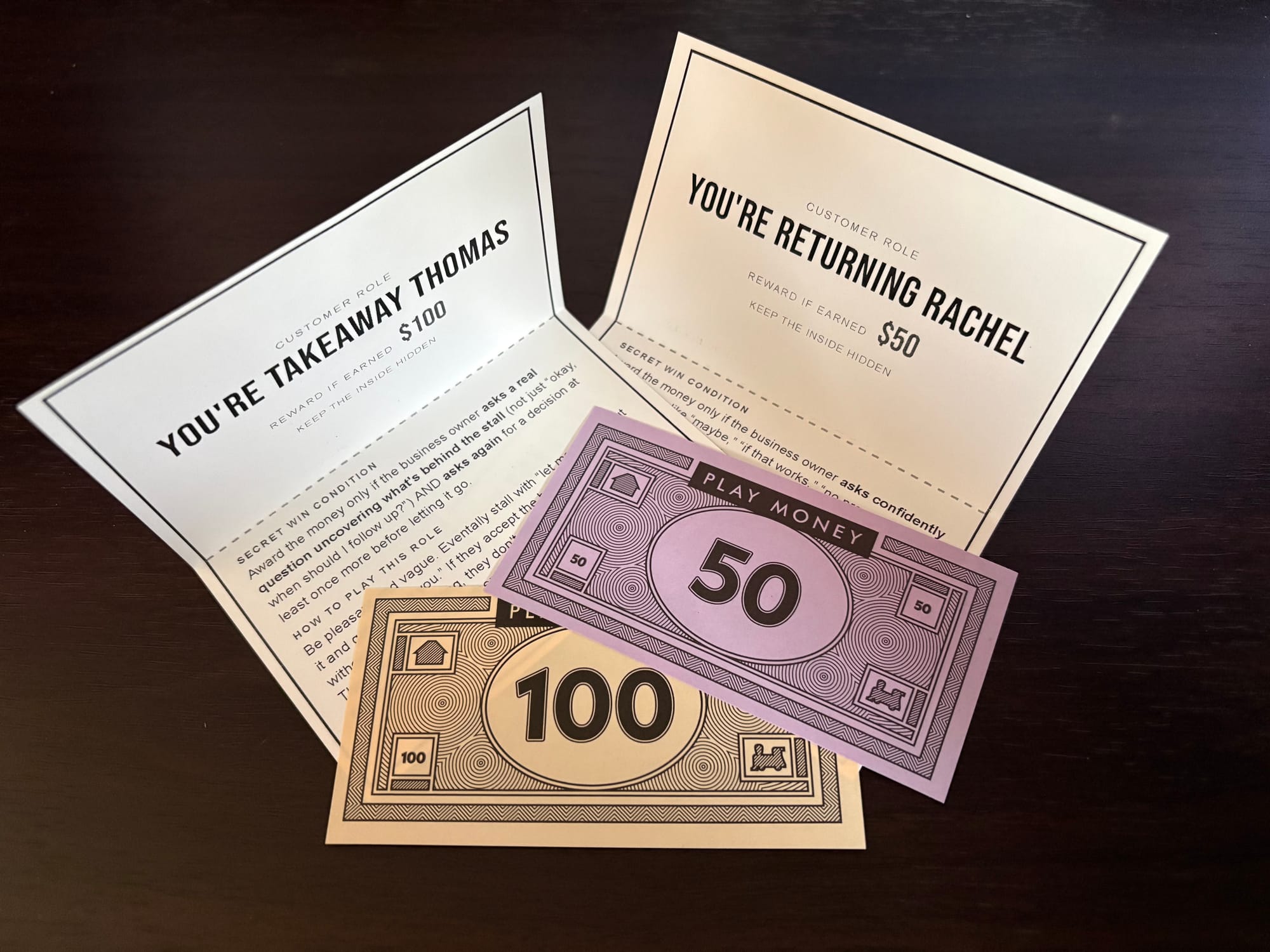 Two role cards for Takeaway Thomas and Returning Rachel sit on a table with 100 dollar and 50 dollar monopoly bills