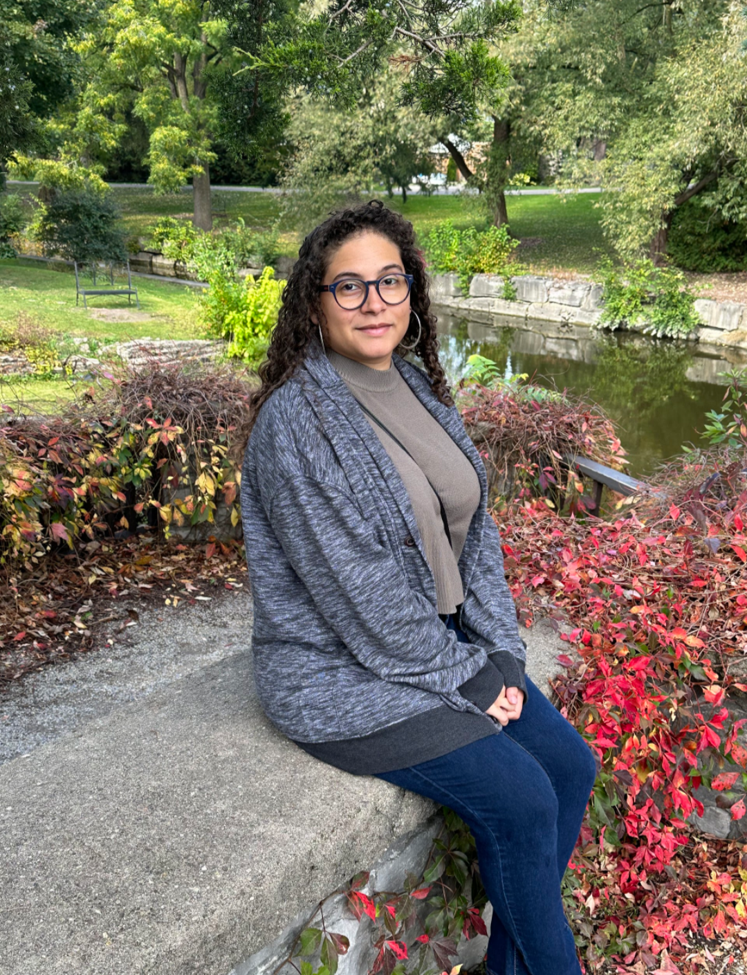 Lissy sits in a park, in front of a river, on a short, concrete wall full of greenery. Lissy's curly brown hair is down and they're wearing a cardigan, glasses, and hoop earrings