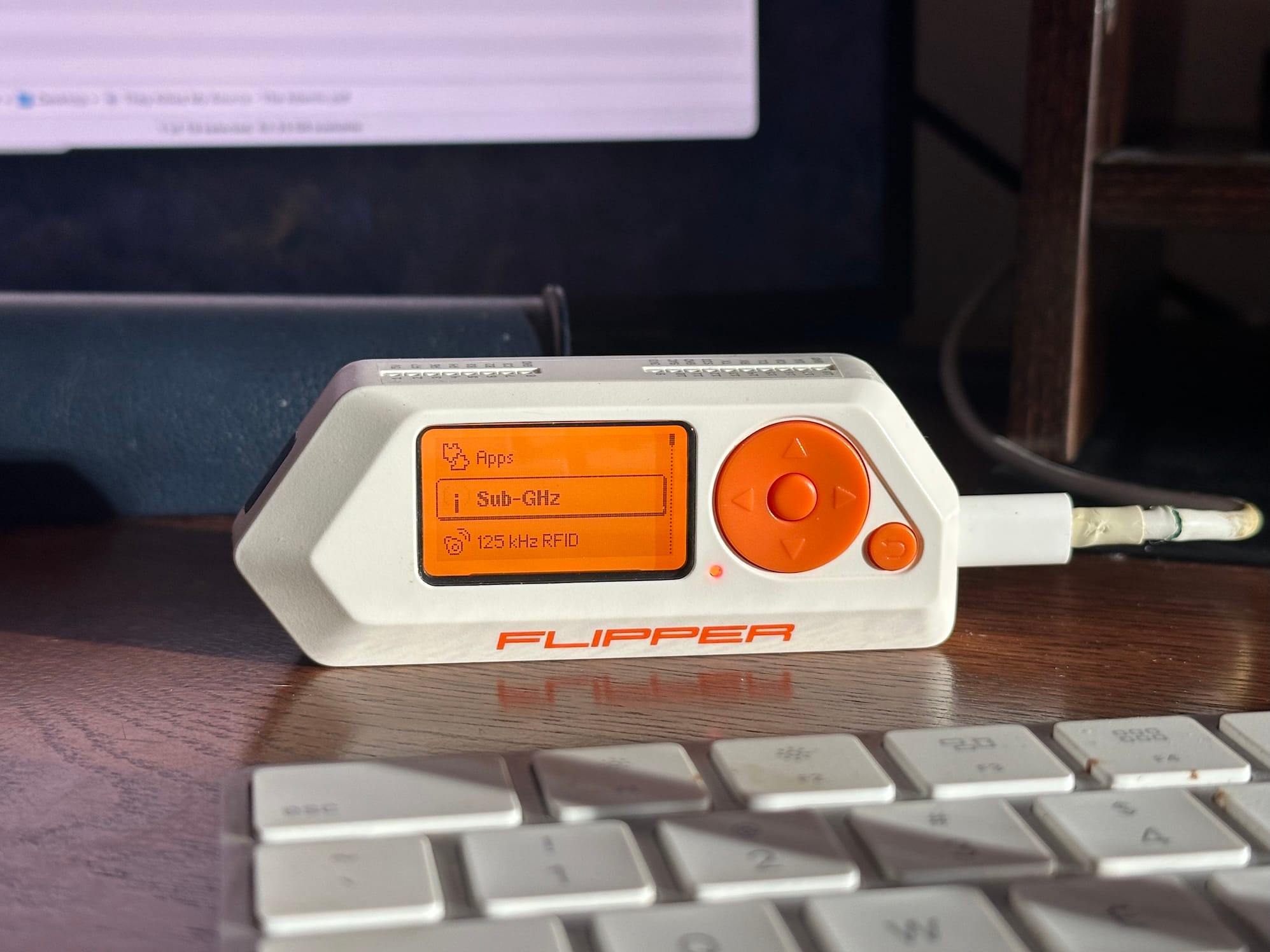 a Flipper Zero tool with its orange display lit up, the device is plugged in and on a wooden table.