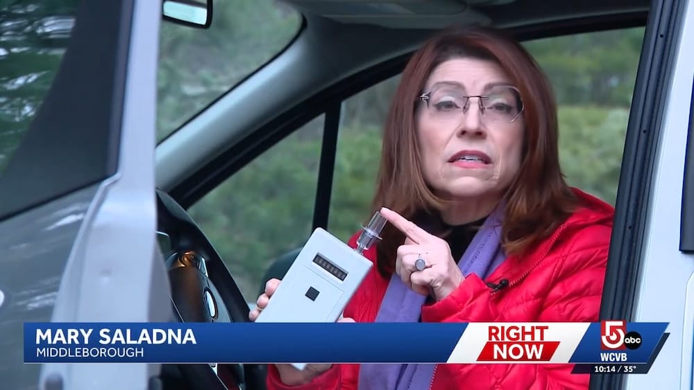 a screenshot of a WCVB video showing reporter Mary Saladna holding one of the Intoxalock breathalyzers while sitting in a car.