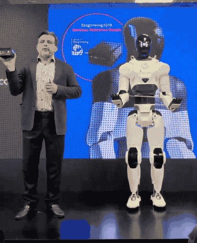 an animated GIF of a humanoid robot suddenly collapsing on stage into itself, next to a person presenting some new technology. in the last few frames, the broken, mangled robot sticks its hand up as if to say "help."