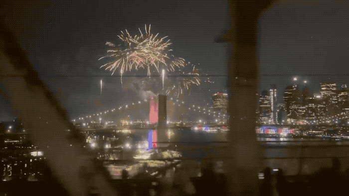 an animated GIF of a view from a moving subway train, going over the Manhattan Bridge between Brooklyn and Manhattan in New York, with fireworks exploding in the sky in the distance.