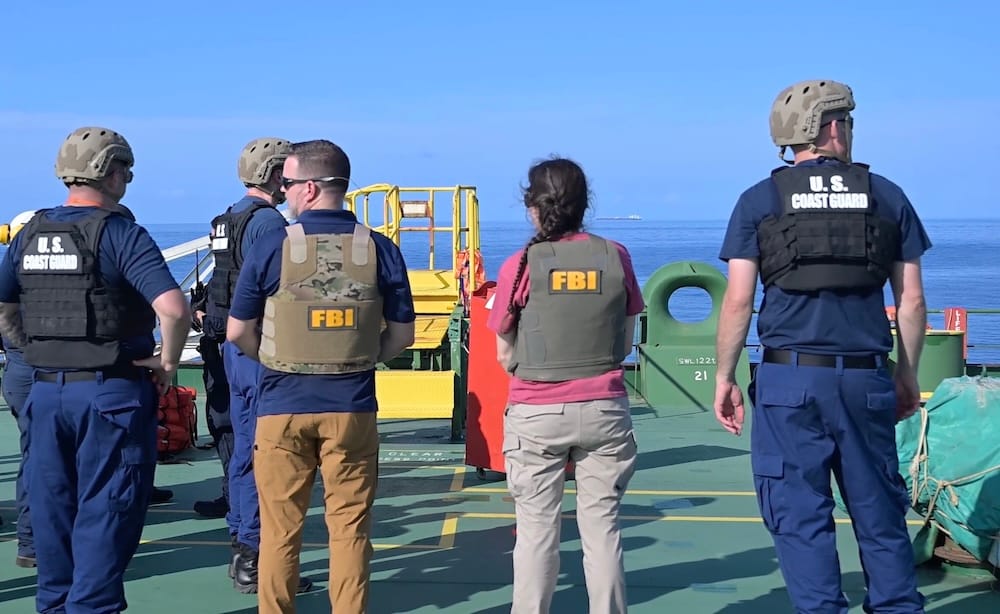 a photo of several U.S. Coast Guard personnel and FBI agents wearing bulletproof vests (for some reason) on a large shipping tanker, looking out to sea, with a big ship in the distance.