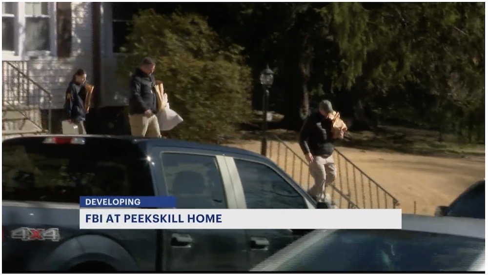 A photo of a live shot of a TV broadcast with FBI agents walking out of a home carrying items, with a chyron saying "FBI AT PEEKSKILL HOME — DEVELOPING"