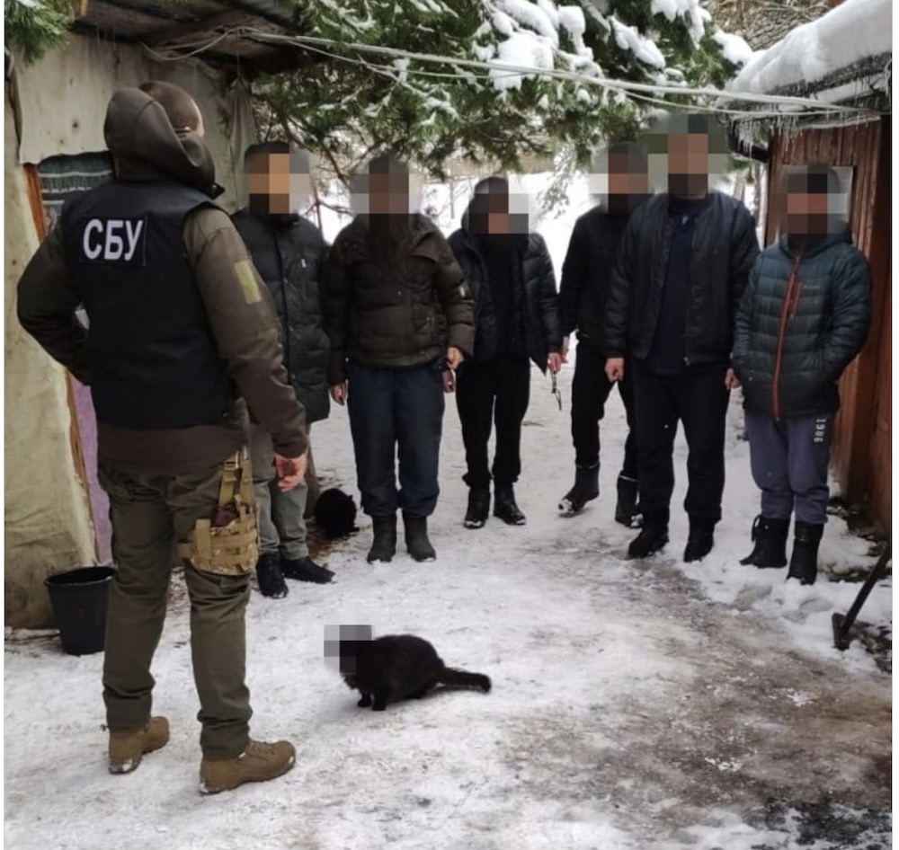A photo of Ukraine police standing with several people outside in the snow with their faces blurred. There's a black cat on the ground, whose face is also blurred.