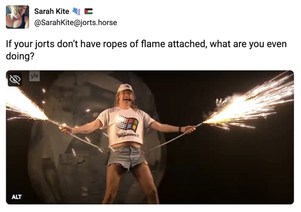 Sarah Kite toot: "If your jorts don’t have ropes of flame attached, what are you even doing?" followed by a photo of Windows95man from Finland on stage at the 2024 Eurovision song competition, singing into a headset microphone, holding his arms out grasping ropes emanating from his jeans shorts, with sparkler flames shooting out of the ends of those ropes (note: he is also wearing a white tshirt and white baseball cap each with the Windows 95 logo