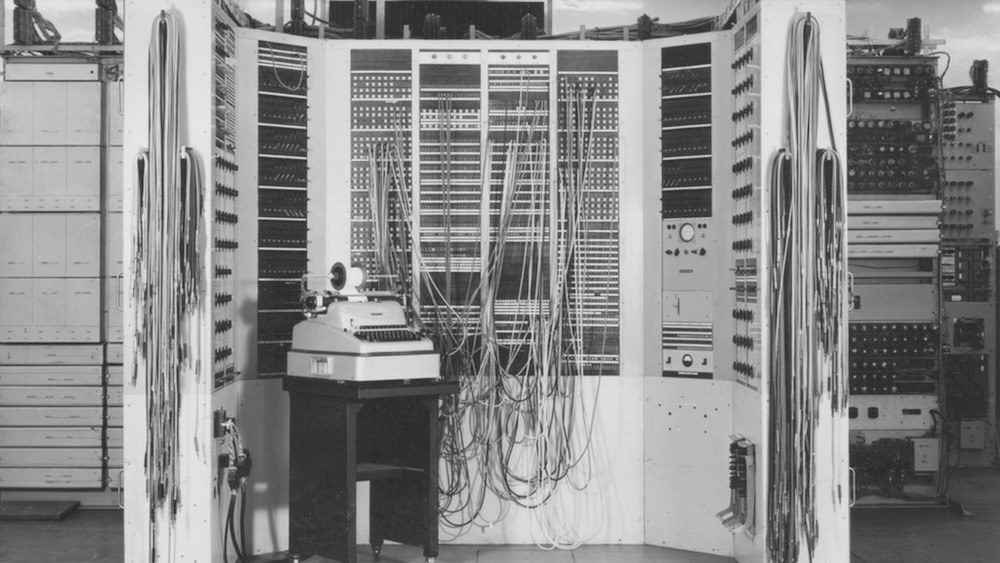 A monochrome photo of the Colossus computer, featuring cables spilling out of a room-sized computer.