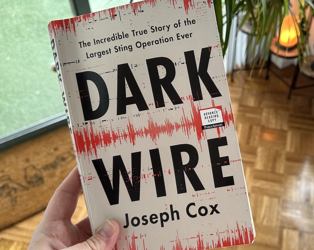 A photo showing my hand holding a copy of Joseph Cox's book, DARK WIRE, which has a audio graphic on the front with the caption, "The Incredible True Story of the Largest Sting Operation Ever."
