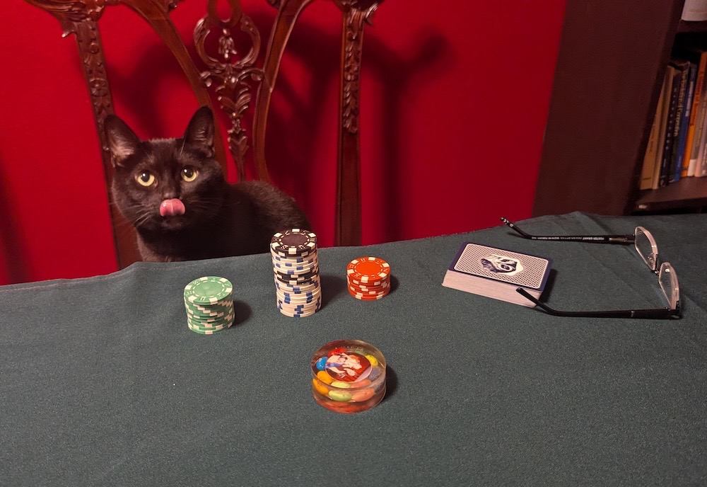 Whisper is a jet-black void floof who is performing an excellent 'mlem' with their tongue, and seen sat on a chair at a poker table with poker chips nearby.