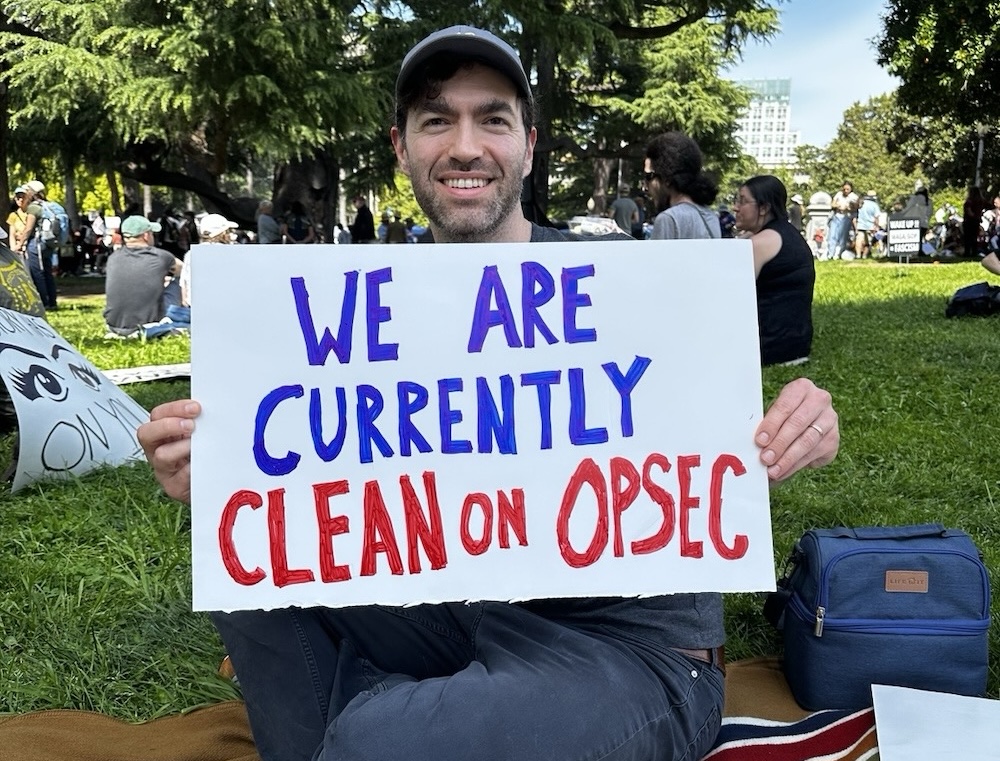 Micah Lee in a park holding a sign that says, “We are currently clean on OPSEC”