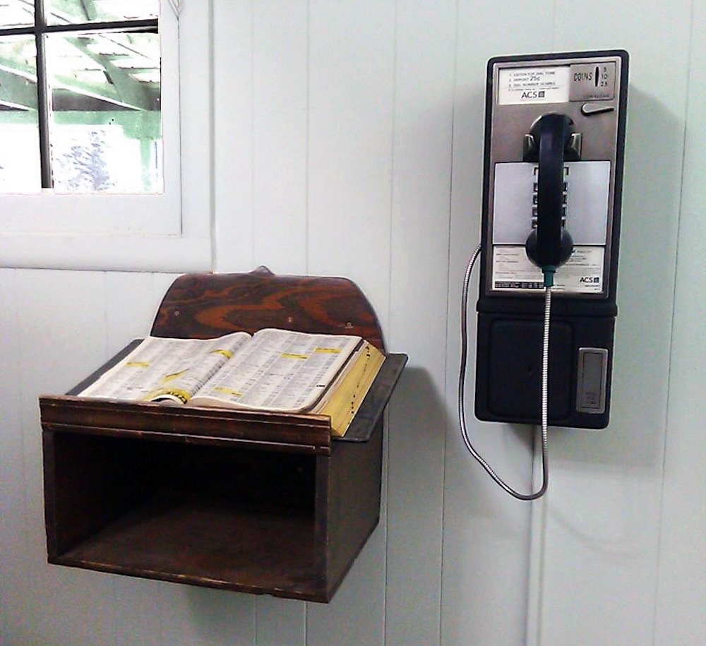 A landline pay-phone on the wooden wall of a building and a yellow pages phone book that contains all the local phone numbers.