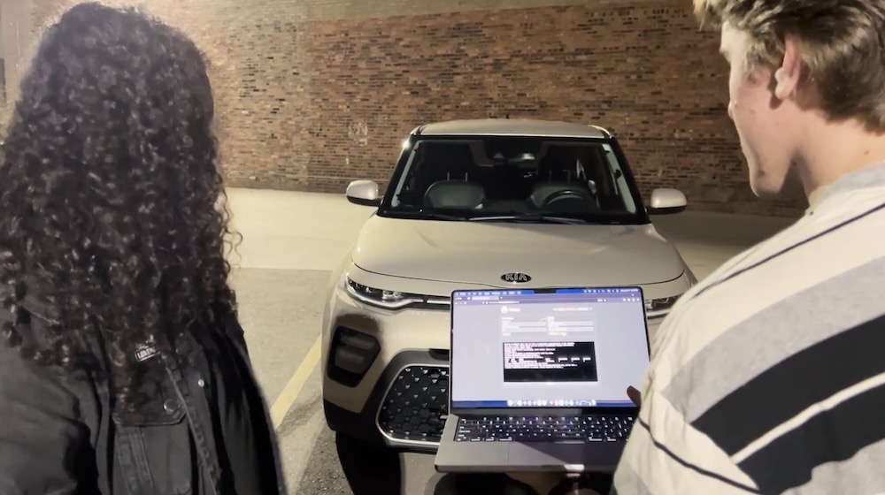 Neiko Rivera (left) and Sam Curry (right), who's holding a laptop running a custom app they made, in front of a Kia car that they're about to remotely hack.
