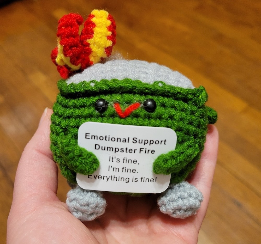 A crocheted dumpster fire holding a sign that says "emotional support dumpster fire. It's fine, I'm fine, everything is fine!" The dumpster is green with a gray top and gray feet, and there's a red and yellow flame on top.