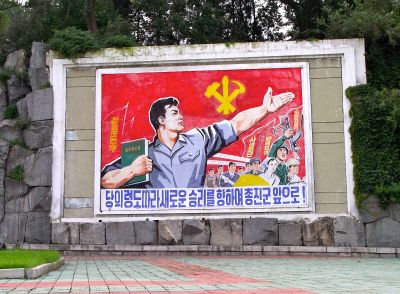 a photo of North Korean propaganda, featuring a man holding a book, gesturing to the North Korean flag, which reads in Korean: "Let's march forward towards a new victory!"