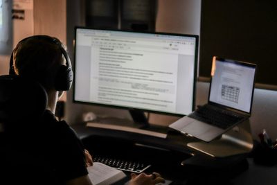 a person sitting facing a computer in a darkened room, with headphones on, reading something on their display.